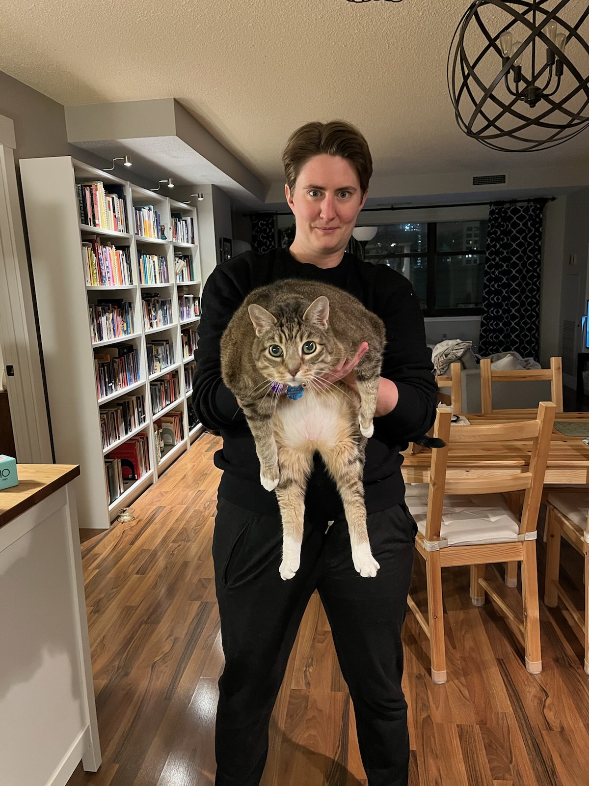 Photo of a person in all black holding a brown tabby cat in front of them in a room with wood floors, with a bookcase and table visible behind them.